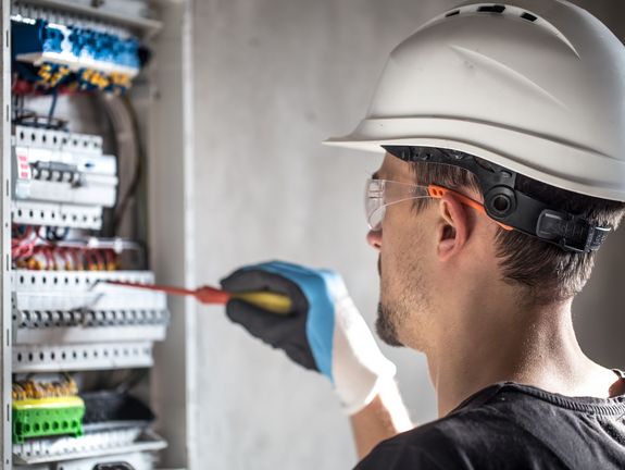 Das Bild zeigt einen Elektriker, der Schutzhelm und Schutzbrille trägt, während er mit einem Schraubendreher an einer offenen elektrischen Schalttafel arbeitet. Die Fokusierung und Konzentration in seiner Haltung verdeutlichen die Sorgfalt und Präzision, die bei dieser Art von Arbeit erforderlich sind. Im Hintergrund sind Kabel und elektrische Bauteile sichtbar, die ein komplexes Netzwerk innerhalb der Schalttafel bilden.