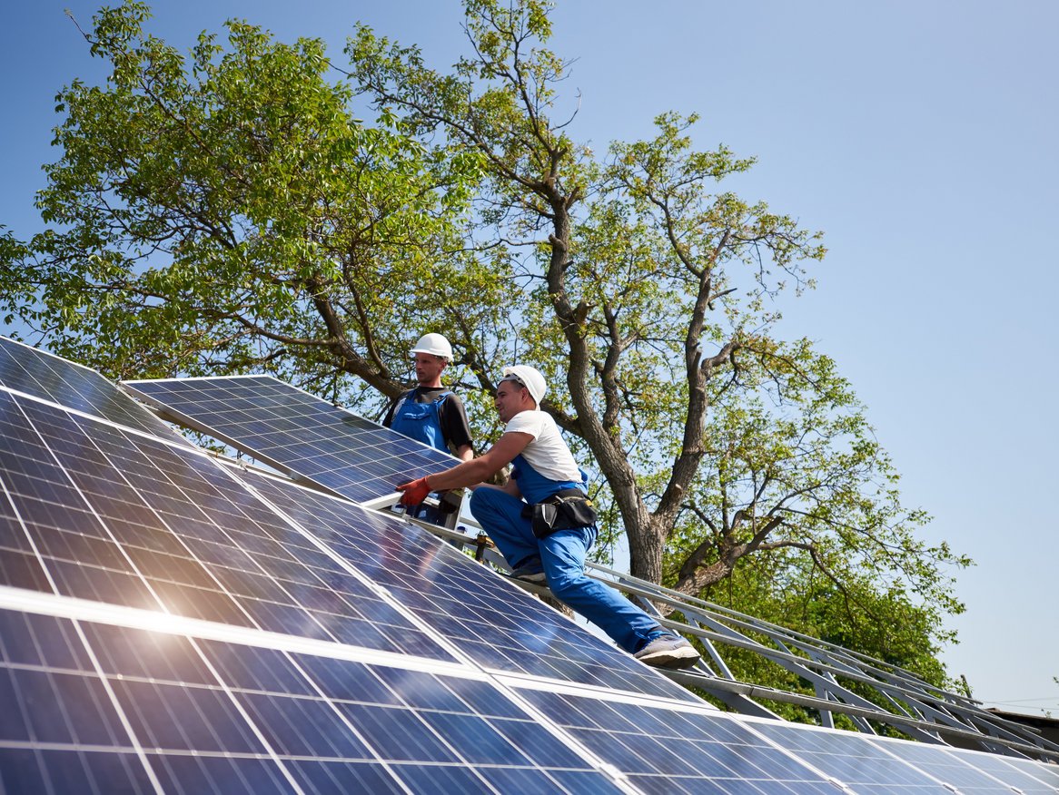Ein Arbeiter montiert Solarpaneele auf einem Hausdach. Die Aufnahme zeigt ihn in Aktion bei strahlendem Sonnenschein, was den Einsatz erneuerbarer Energien in Wohngebieten hervorhebt.
