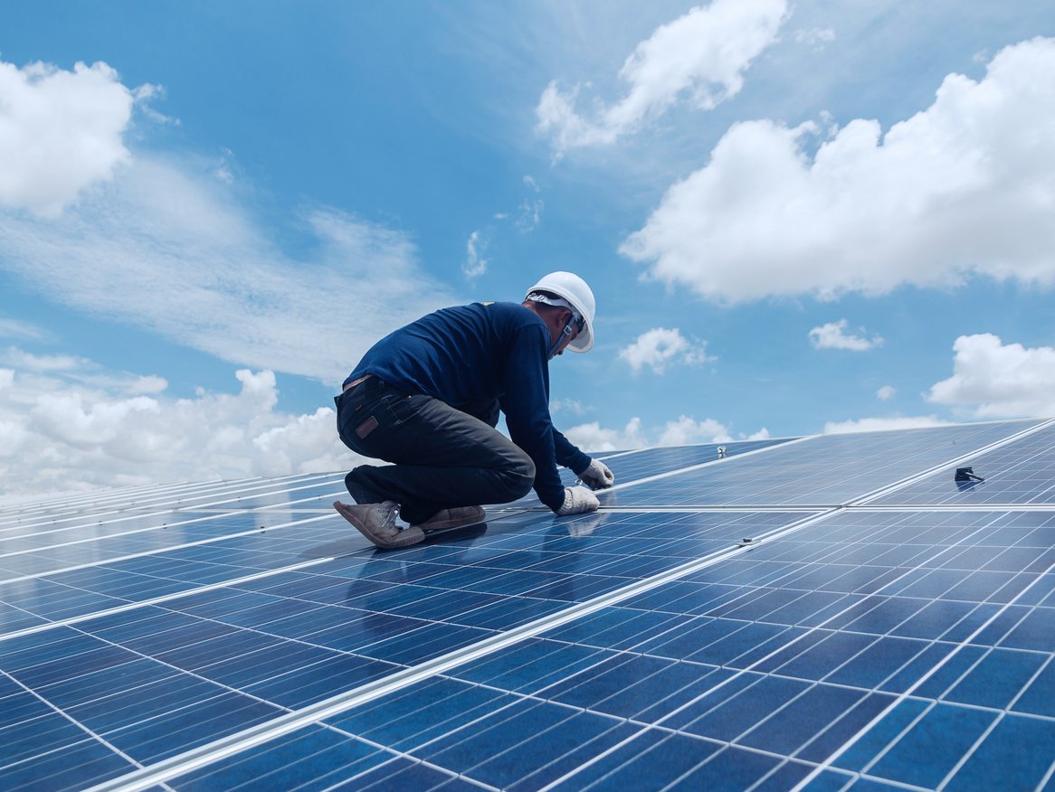 Ein Techniker in Arbeitskleidung installiert Solarpanels auf einem großen Solarkraftwerk unter einem blauen Himmel mit weißen Wolken, was die Verbindung von menschlicher Arbeit und erneuerbaren Energien veranschaulicht.