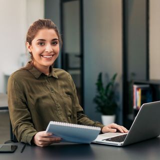 Eine Frau mit einem freundlichen Lächeln sitzt an ihrem Arbeitsplatz vor einem Laptop und hält Dokumente in der Hand. Ihre professionelle Haltung und das moderne Büro im Hintergrund spiegeln eine angenehme Arbeitsumgebung wider.