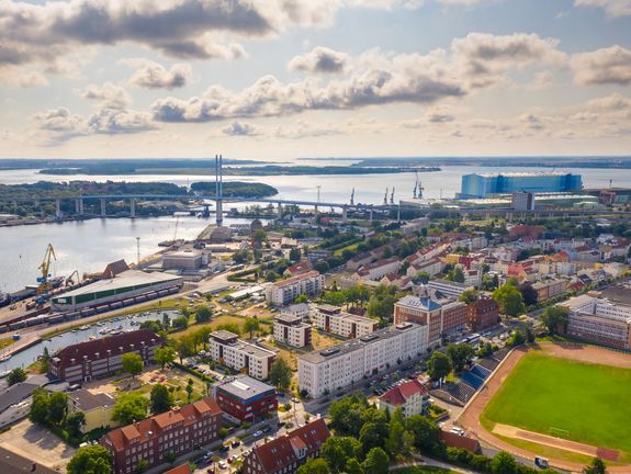 Ein Luftbild zeigt eine Stadt am Wasser, mit einer Brücke, die zwei Ufer verbindet, und industriellen Strukturen im Hintergrund. Die Mischung aus Wohnvierteln, Gewässern und industriellen Anlagen gibt einen Eindruck von der Vielfalt städtischer Umgebungen.