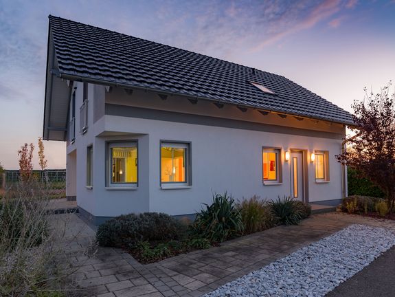 Gemütliches Einfamilienhaus bei Dämmerung mit beleuchteten Fenstern und einem markanten schwarz-weißen Dach, umgeben von Bäumen und einem kiesbedeckten Vorgarten.