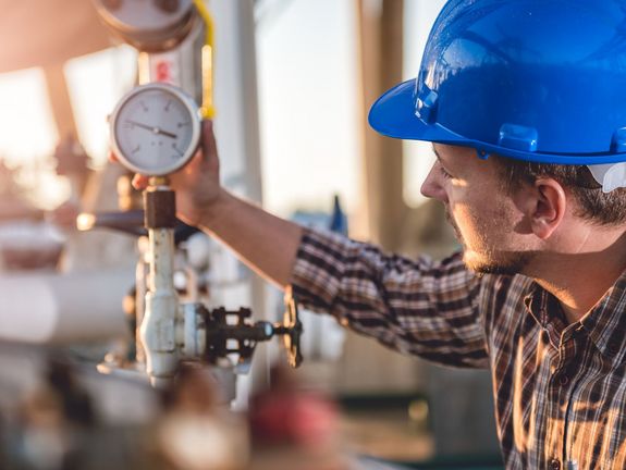 Ein Ingenieur in einem blauen Schutzhelm überprüft aufmerksam ein Manometer in einer industriellen Anlage. Seine Konzentration und die professionelle Ausrüstung deuten auf wichtige Wartungsarbeiten oder Qualitätskontrollen in einem technischen Umfeld hin.