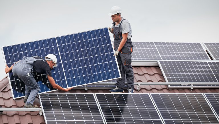 Zwei Monteure in Arbeitskleidung und Schutzhelmen installieren sorgfältig Solarpaneele auf einem Hausdach. Diese Szene hebt das Engagement für erneuerbare Energien und die Wichtigkeit der Solarenergie für eine nachhaltige Zukunft hervor.