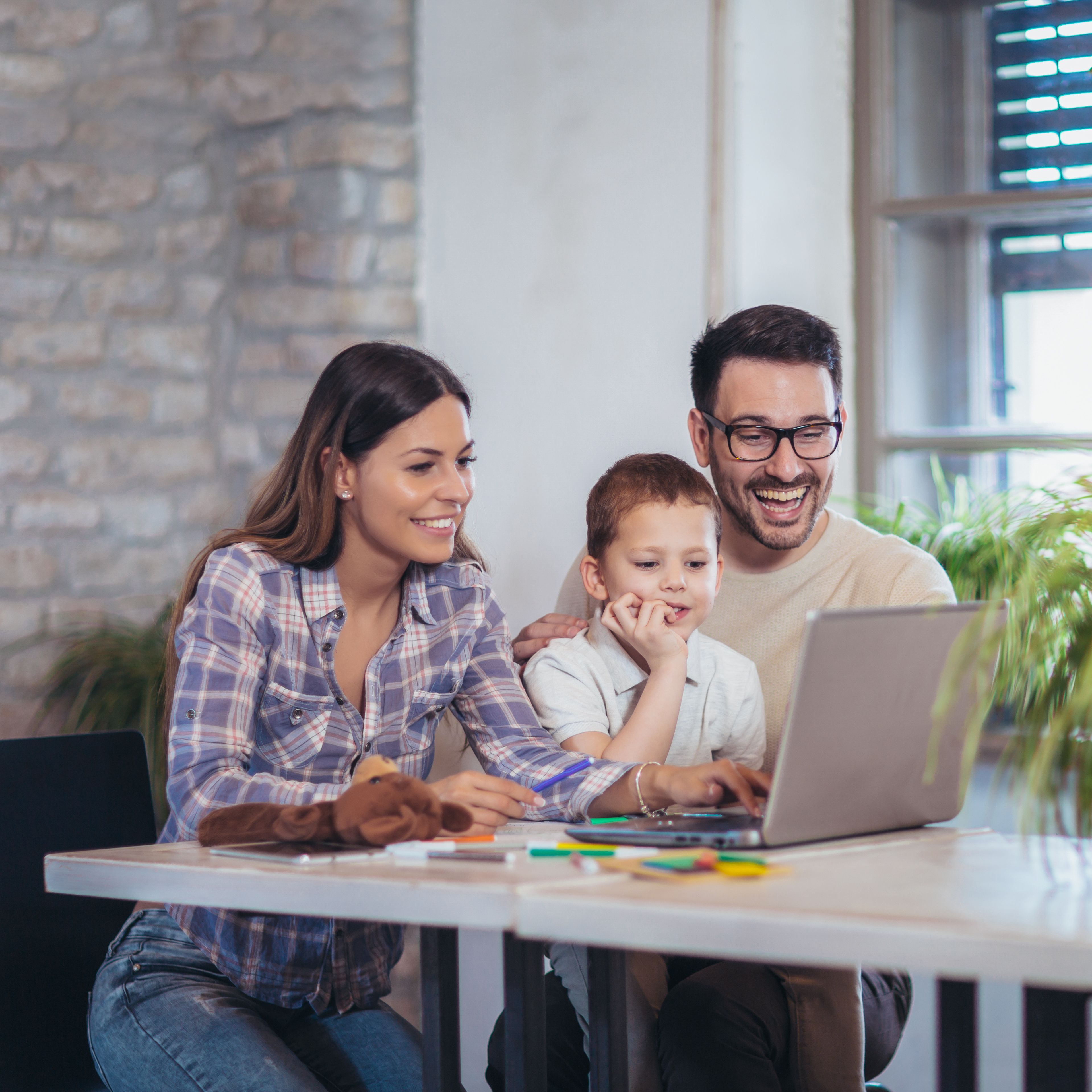 Das Bild zeigt eine Familie mit einer Tochter, die gemeinsam am Esstisch sitzt und auf einen Laptop schaut. Sie befinden sich in einem hellen Raum mit einer Ziegelwand und Fenstern, durch die Tageslicht einfällt.