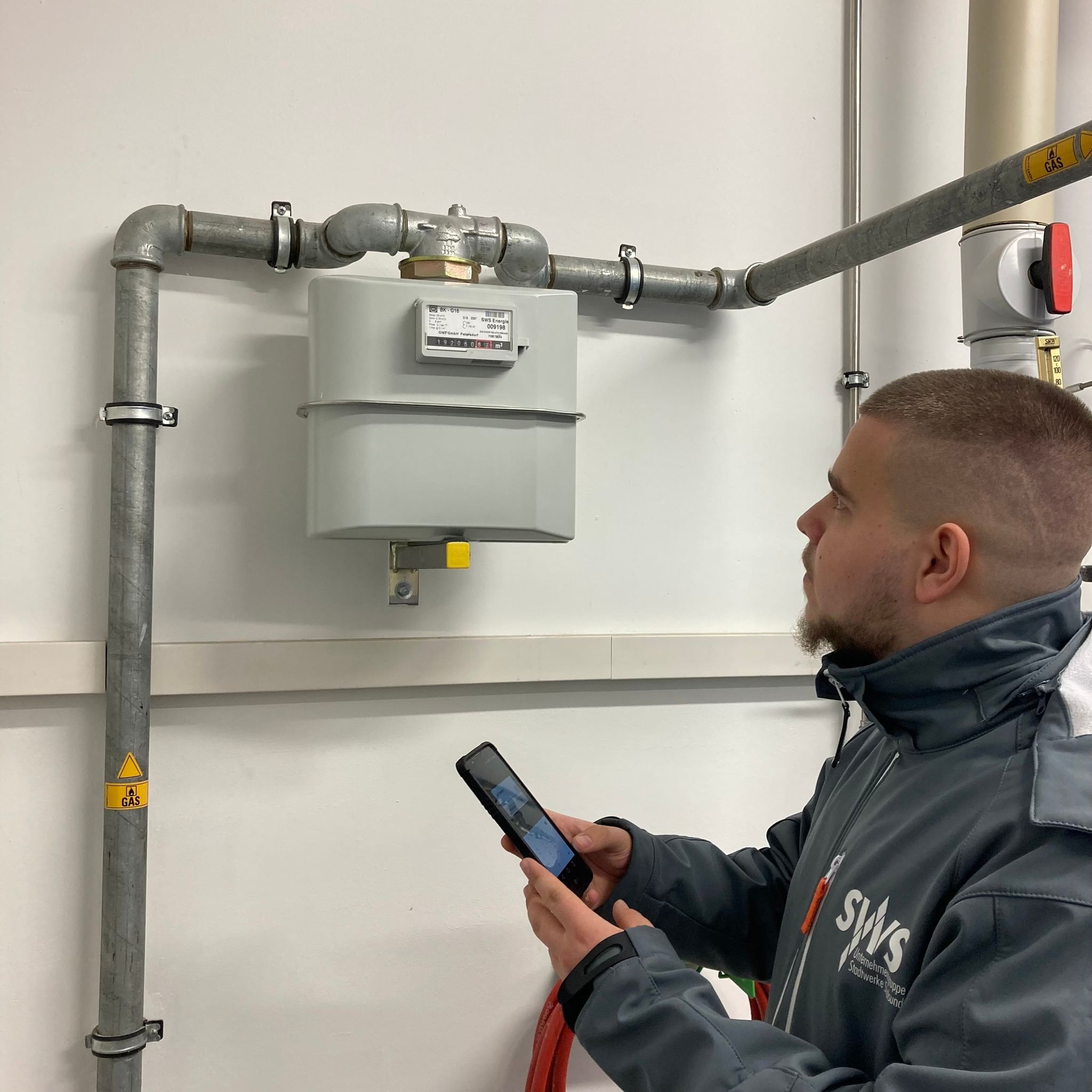 Ein Zählerableser der Stadtwerke Stralsund prüft aufmerksam einen Gaszähler und hält ein mobiles Erfassungsgerät in der Hand in einem technischen Raum.
